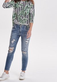 Camisa de rayas en blanco y negro con un patrón de hojas verdes, combinada con unos pantalones skinny rasgados en azul claro y zapatillas blancas.