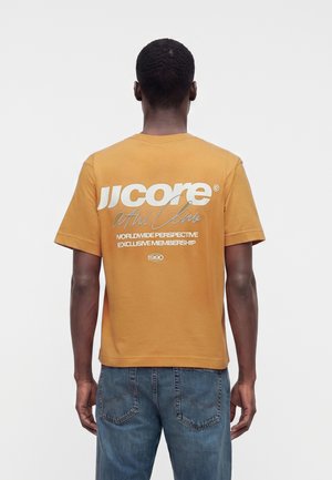 Homme de dos, portant un t-shirt jaune moutarde avec texte blanc et gris et un jean bleu, sur un fond clair uni.