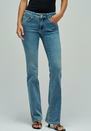 Femme portant un jean droit bleu clair et des sandales noires à talons et brides, debout avec une main dans la poche devant un fond uni.