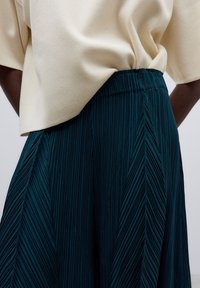 Un gros plan d'une personne portant un haut beige et une jupe plissée vert foncé, mettant en valeur la texture et le design du tissu.