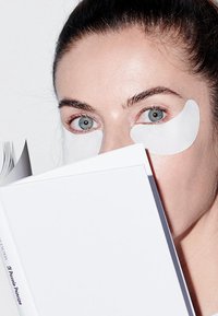 Patch idratanti per gli occhi sulla pelle, con un libro bianco parzialmente visibile e con un dorso scuro. Sfondo bianco uniforme.