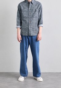 Blauwe geblokte button-up shirt met opgerolde mouwen, gecombineerd met losse blauwe denim jeans en witte sneakers. Platte voeten, effen achtergrond.