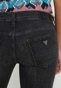 Jeans de mezclilla negra con un ajuste slim, que presentan un pequeño parche de logo triangular, bolsillos traseros y detalles de costura sutiles.