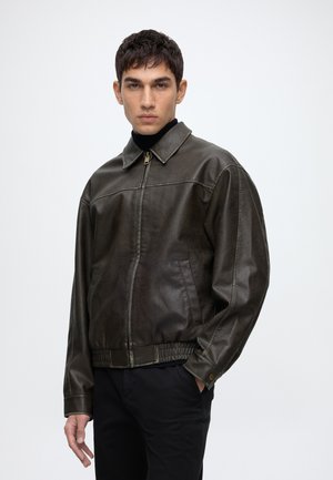Jeune homme portant une veste en cuir marron foncé sur un col roulé noir avec un pantalon noir, debout devant un fond clair uni.