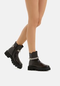 Stivaletti neri alla caviglia con finitura lucida, suola in gomma spessa e una catena argentata decorativa attorno al polsino. Presentano una cerniera laterale per la chiusura.