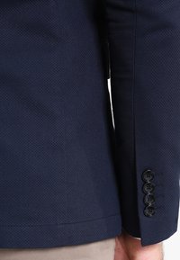 Marinblå blazer med en strukturerad yta, med rak skärning, fyra svarta knappar på ärmarna och en ren fåll.