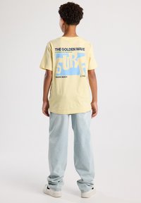 Lichtgele katoenen t-shirt met een groot grafisch ontwerp op de achterkant met blauwe en zwarte tekst. Gecombineerd met losse lichtblauwe jeans.