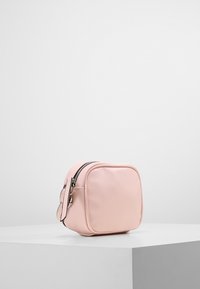 Piccola borsa quadrata in pelle rosa chiaro con zip e manico da polso, posizionata su un pedestal bianco contro uno sfondo chiaro e semplice.