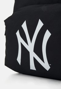 Sort rygsæk med et stort, hvidt NY-logo. Fremstillet af slidstærkt stof, har den en glat tekstur og diskrete syninger.