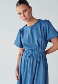 Modra denim obleka s kratkimi puf rokavi, elastičnim pasom in tekočo krilo. Gladka tekstura z rahlim leskom.