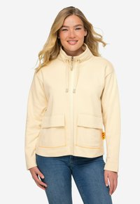 Chaqueta beige con cremallera y cuello alto, dos bolsillos frontales, etiqueta de acento naranja y puños de canalé. Tejido suave con una textura lisa.