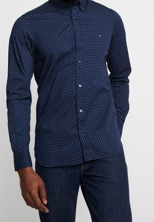 Man draagt een marineblauw overhemd met kleine witte geometrische patronen en donkere blauwe spijkerbroek, staand tegen een effen achtergrond.