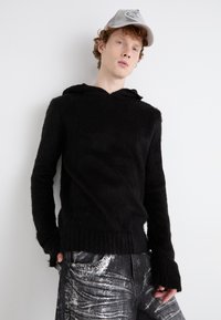 Moleton preto felpudo com um corte solto, apresentando capuz e mangas longas, combinado com jeans cinza desgastados decorados com padrões.