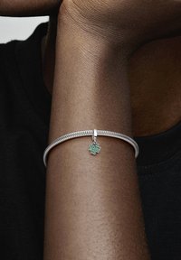 Srebrn pleteni zapestnica z zeleni emajliranim pravečim deteljico, prikazana na temnopolti roki, proti črnemu ozadju.