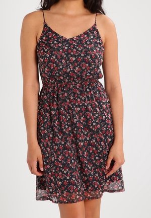 Femme portant une robe noire sans manches avec un motif floral rouge et blanc et des bretelles fines, se tenant devant un fond uni.