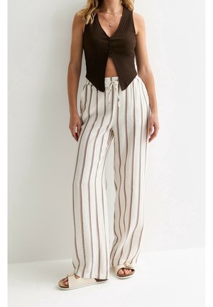 Femme portant un haut sans manches marron côtelé à boutons, un pantalon blanc à jambes larges avec des rayures verticales marron, et des sandales beiges ouvertes à l'avant.