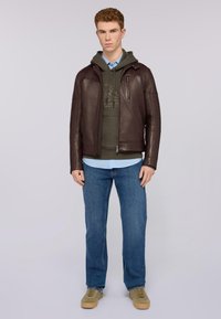 Brązowa skórzana kurtka z przednim zamkiem, połączona z zieloną bluzą z kapturem z logo, jasnoniebieską koszulą i niebieskimi dżinsami. Beżowe tenisówki.