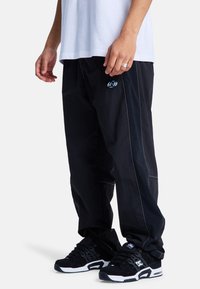 Black nylon pants with white stitching and a small logo on the pocket, paired with black sneakers featuring white soles.