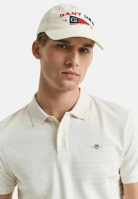 Polo crème à rayures subtiles, doté d'un plastron à deux boutons et d'un logo Gant sur la poitrine. À associer avec une casquette blanche ornée de graphismes rouges et navy.