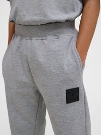 Person iført grå træningbukser med elastisk talje, hænder i lommerne og sort rektangulært logo-patch på låret.