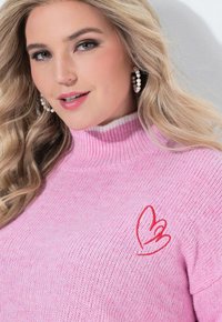Roze gebreide trui met een hoge ribboordkraag, met subtiele textuur en een rood hartontwerp aan de linkerkant.