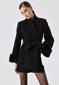 Cappotto nero avvolgente con vita cintata, polsini con pelliccia e colletto strutturato. Tessuto testurizzato in una silhouette sartoriale.
