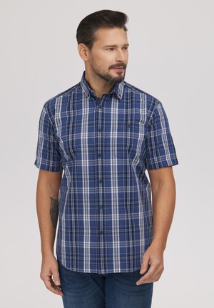 Mężczyzna o krótkich ciemnych włosach i brodzie, ubrany w niebieską i białą kraciastą koszulę na krótki rękaw z guzikami, stojący na jednolitym tle.