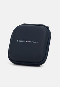 Tamsiai mėlynas tvirtas dėklas su lygiu paviršiumi, turintis užtrauktuko uždarymą ir baltą "TOMMY HILFIGER" logotipą, esantį centrinėje priekio dalyje.