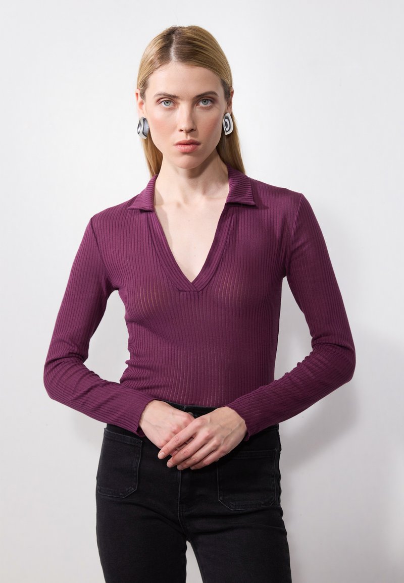 Long-sleeve, deep V-neck top in ribbed burgundy fabric. Features a collared neckline and fitted silhouette, paired with dark jeans.