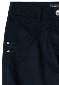 Primer plano de pantalones negros con bolsillo delantero, trabilla para cinturón, botón plateado y detalles de remaches en la tela y costuras.