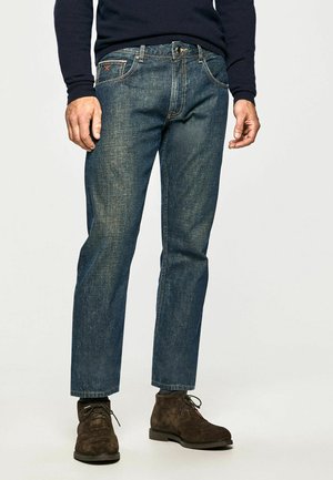 Homme portant un jean bleu délavé, un pull bleu marine foncé et des bottines en daim marron foncé à lacets, debout sur un fond clair uni.