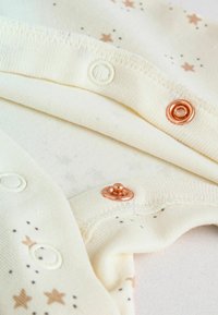 Tissu de couleur crème avec des motifs d'étoiles, présentant des boutons-pression de couleur cuivrée et une texture douce. Le design comprend des bords côtelés et des accents ludiques.
