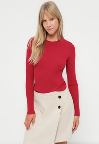 Rød, ribbestrikket langermet topp kombinert med en lys beige omslagskjørt med svarte knapper og en sidelomme. Glatt, tettsittende tekstur.