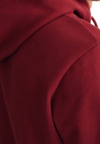 Close-up van een persoon in een donkerrode hoodie, met de nadruk op de schouder en de details van de aan de hoodie bevestigde kap.