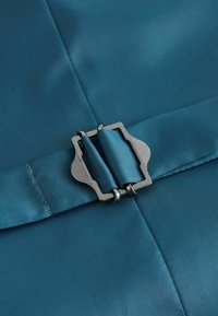 Detailní záběr na stříbrnou kovovou přezku, která upíná sklopený pásek z tyrkysově modrého saténu na odpovídajícím tyrkysově modrém oděvu.