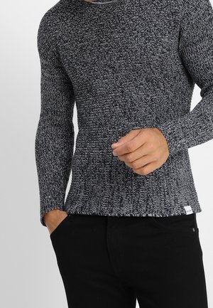 Mężczyzna noszący dopasowany, szary i czarny melanżowy sweter z długim rękawem, jedną ręką w kieszeni, a drugą poprawiający rękaw.