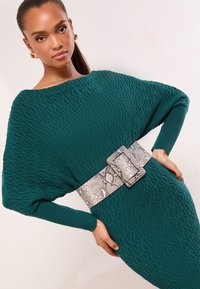 Vestido de punto color verde azulado con textura geométrica, de corte extragrande y mangas largas, ceñido en la cintura con un cinturón plateado con estampado de piel de serpiente.