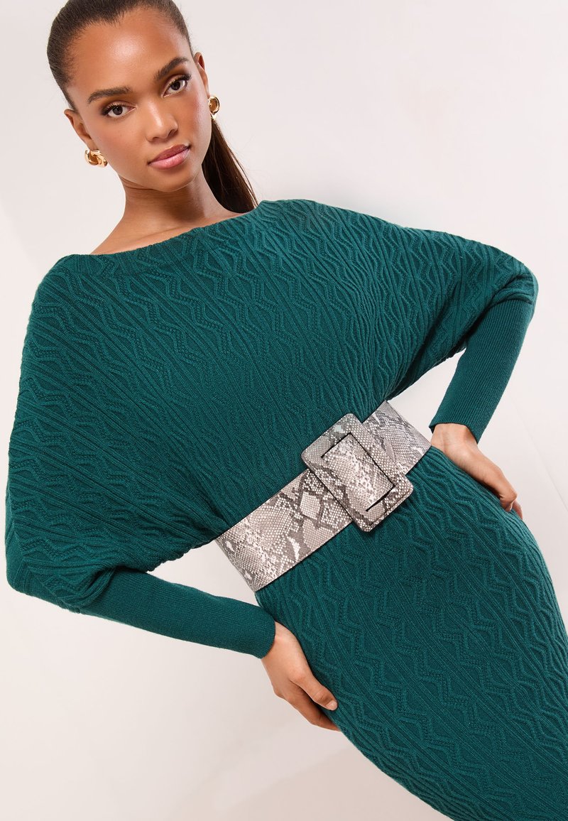 Vestido de punto color verde azulado con textura geométrica, de corte extragrande y mangas largas, ceñido en la cintura con un cinturón plateado con estampado de piel de serpiente.