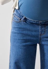 Jeans de ganga azuis com corte reto, apresentando bolsos cosidos e um fecho de botão clássico. A textura é suave com um desbotamento subtil.