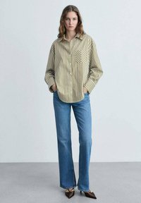 Camicia button-up a righe in oliva e crema, con taschino sul petto. Abbinata a jeans azzurri chiari, leggermente svasati, e scarpe marroni a punta.