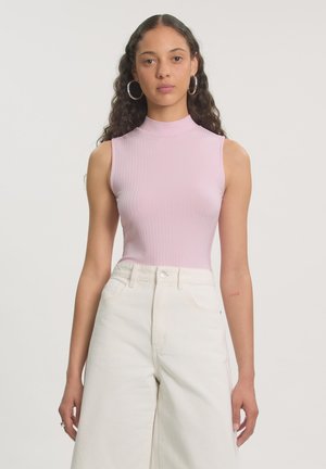 Jonge vrouw met lang krullend haar, met een mouwloos roze geribd topje en een witte broek met hoge taille, staand voor een effen achtergrond.