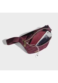 Maroon nylon fanny pack with a gold zipper and logo. Interior contains a phone and earbuds. Features adjustable strap and textured exterior.