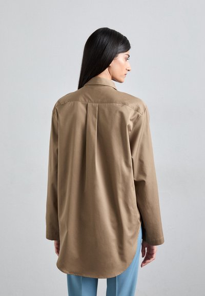 Brown long-sleeve shirt with a collar, featuring a straight back yoke and a rounded hem, paired with light blue pants.