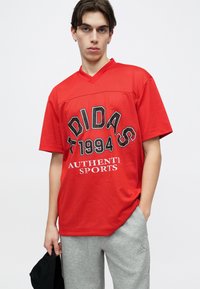 Νέος άνδρας που φοράει κόκκινη αθλητική μπλούζα Adidas με το κείμενο «1994 Authentic Sports», γκρι αθλητική φόρμα Adidas, κρατώντας ένα μαύρο καπέλο, σε λευκό φόντο.