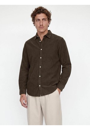 Homme aux cheveux bouclés et à la moustache, portant une chemise marron foncé boutonnée et un pantalon beige clair, debout avec une main dans la poche.