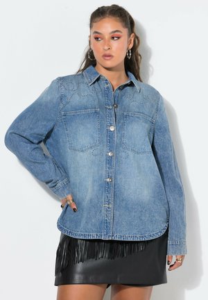 Kvinde iført en blå denimjakke med knapper og forlommer og en sort lædernederdel med frynser, poserer med hånden på hoften.