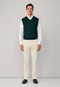 Zielony sweter w kształcie litery V na białej koszuli, w połączeniu z kremowymi spodniami i czarnymi butami. Prosty, klasyczny krój i gładka faktura.