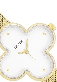 Montre avec bracelet en maille dorée et cadran blanc en forme de trèfle, avec des aiguilles des heures et des minutes dorées et un logo noir. Verre clair.