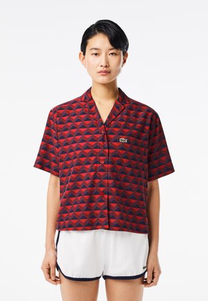 Camisa de manga corta con un patrón geométrico en rojo y azul marino, hecha de tela. Presenta un cuello, cierre de botones en el frente y un logo sutil en el pecho.