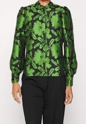 Kvinde iført en sort bluse med stor grøn blomsterbroderi og puffede ærmer, kombineret med sorte bukser.